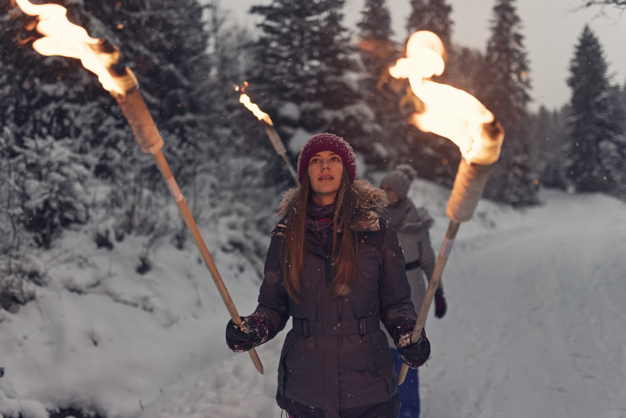 Ein Mädchen im Teenageralter geht einen schneebedeckten Waldweg entlang, während sie zwei brennende Fackeln hält, eine in jedem Handschuh. Sie trägt einen braunen Knit-Hut und eine dunkle Winterjacke. Ihre Mutter folgt dem Mädchen mit einer weiteren Fackel. Die Bäume sind mit Schnee bedeckt und das Licht ist gedimmt. Show mit Canon R5 an einem dunklen Winterabend.